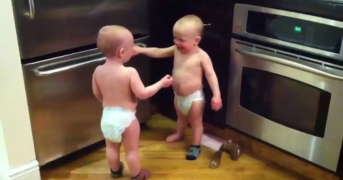 Two toddlers, one in a diaper, interact playfully in a kitchen with stainless steel appliances.