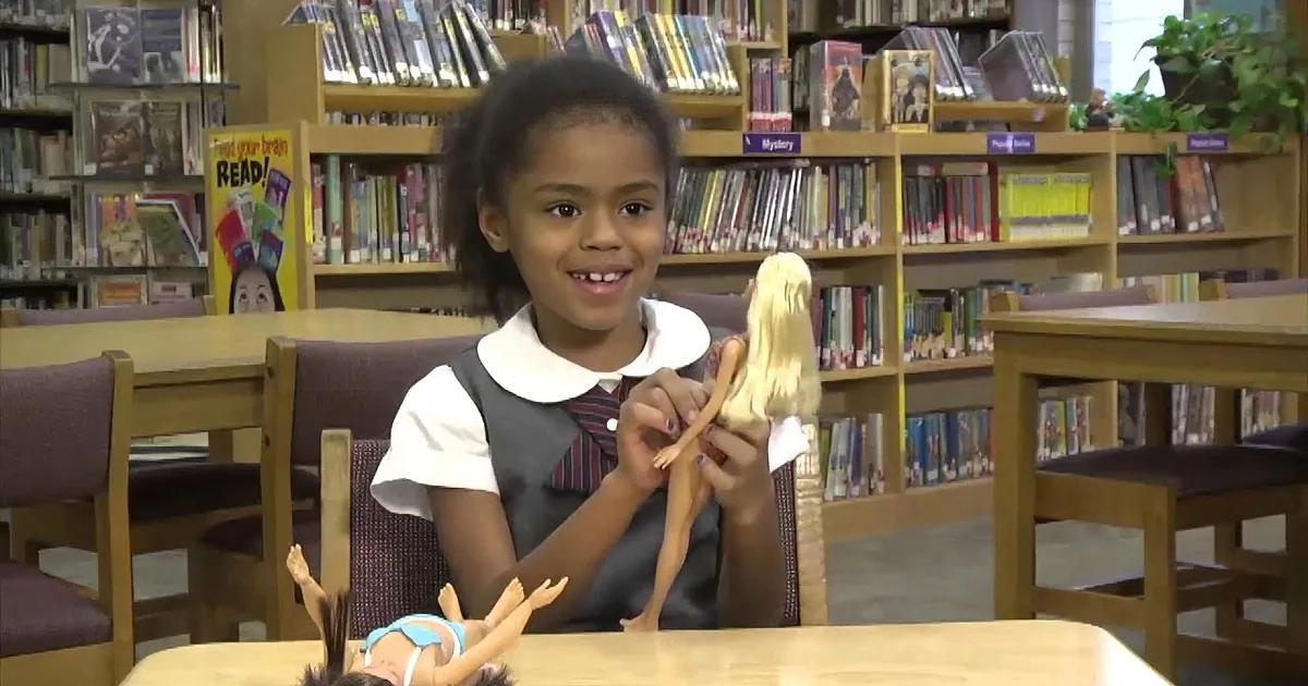 A young girl smiles while holding a doll in a library setting with bookshelves in the background.