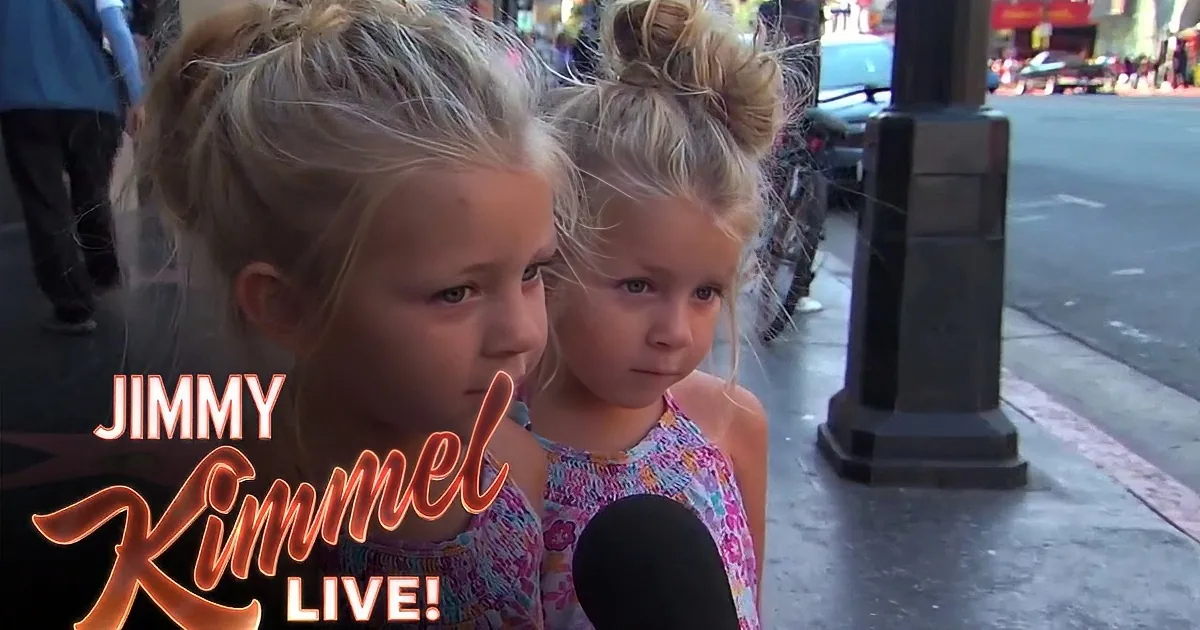 Two young girls with blonde hair in buns are being interviewed on a city street.