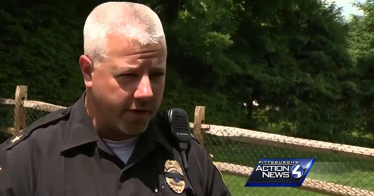 A police officer in uniform stands outdoors, speaking, with a fence and trees in the background.
