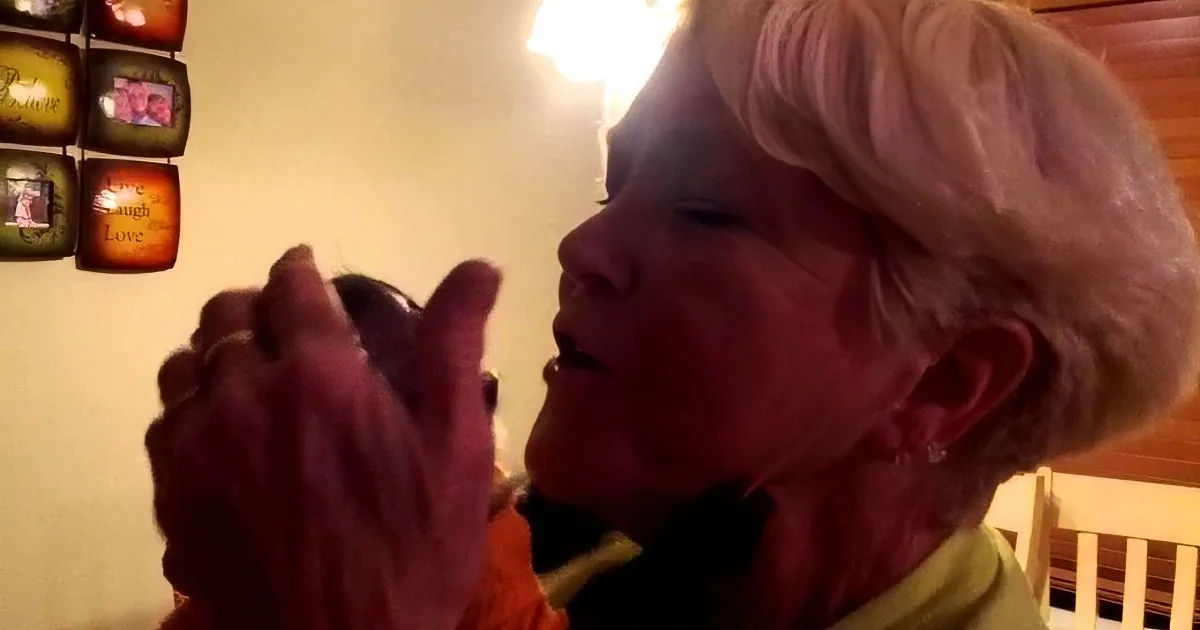 An elderly woman with short gray hair is holding and examining a small object, possibly a piece of jewelry or a small tool.