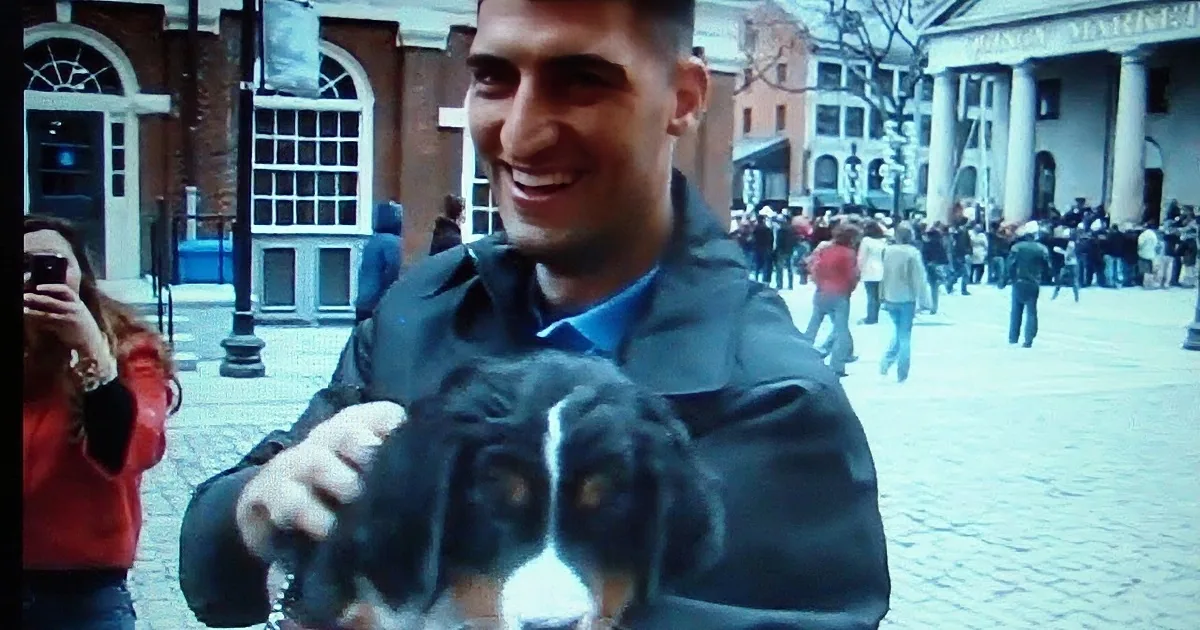 A man smiles while holding a dog in a bustling outdoor area with people and buildings in the background.