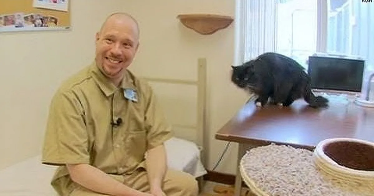 A man in a tan shirt smiles while a black cat sits on a table in the background.