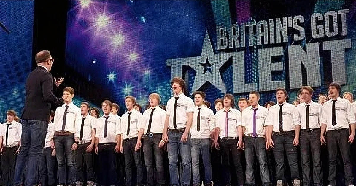A man in a suit stands on stage with a choir in white shirts and ties, preparing for a performance on "Britain's Got Talent."