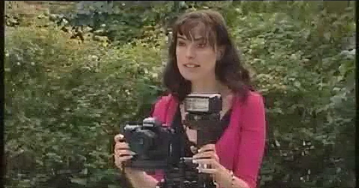 A woman in a pink top holds a camera with a flash, surrounded by green foliage.