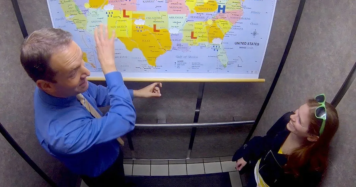 A man in a blue shirt gestures towards a map of the United States while a woman in a black jacket and sunglasses looks up...