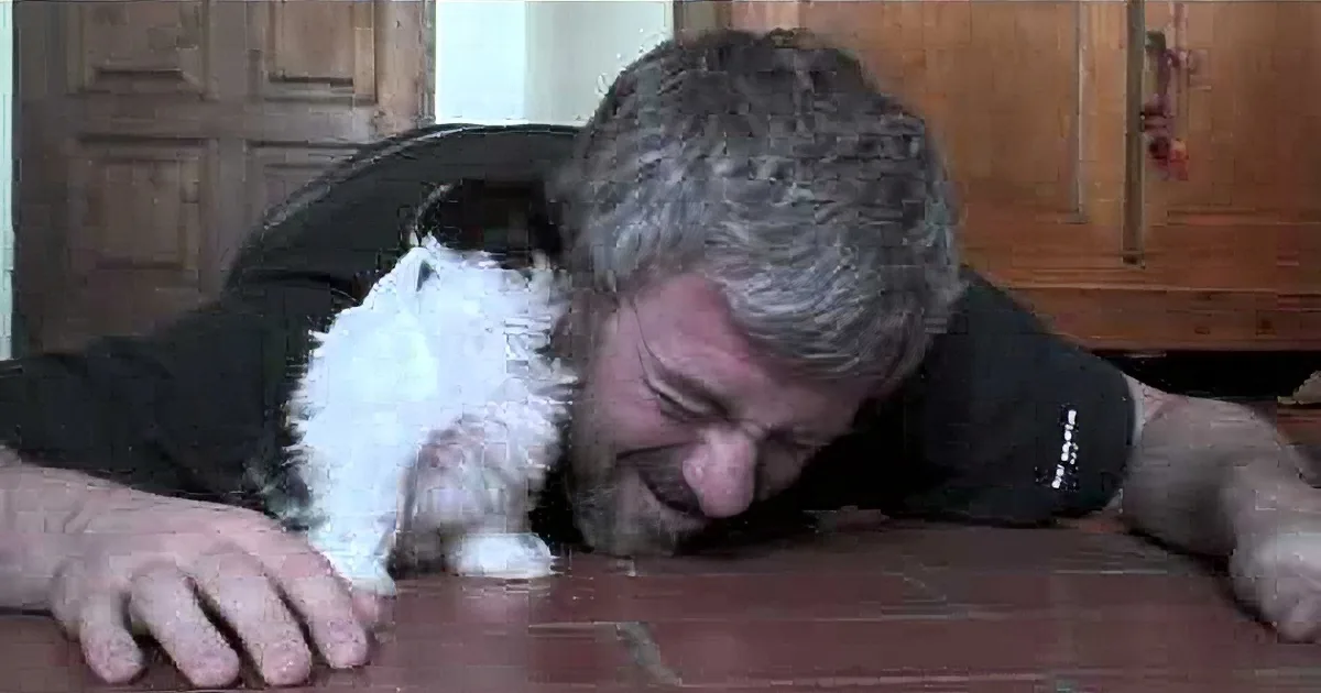 A man is playfully interacting with a small black and white kitten on a tiled floor.