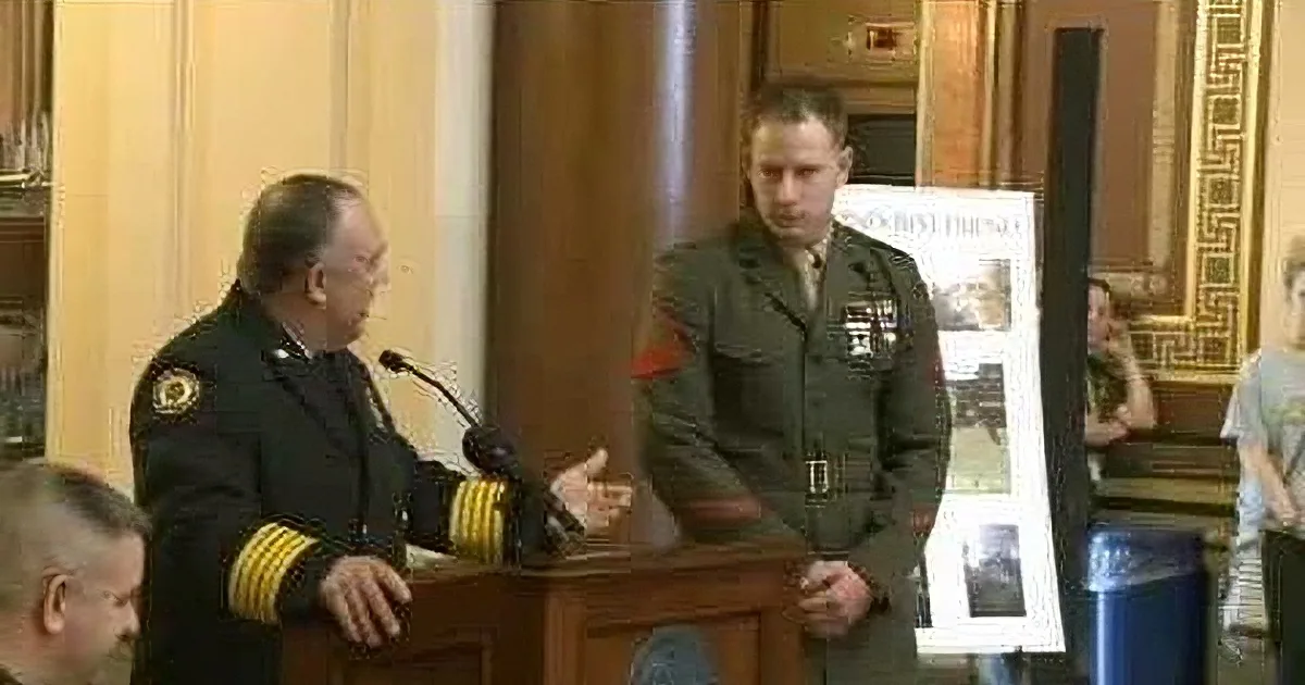 A man in a navy uniform speaks at a podium, gesturing to another man in a military uniform standing beside him.