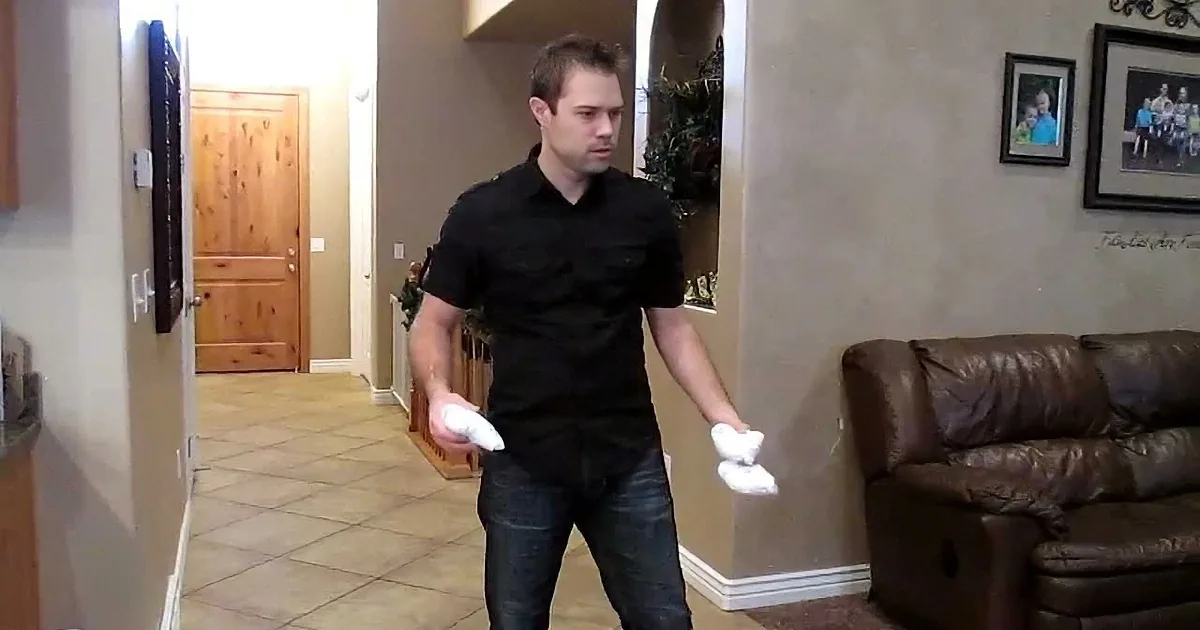 A man in a black shirt and jeans holds white objects, possibly controllers, in a living room setting.