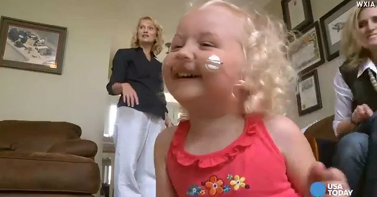 A young girl with curly blonde hair and a pink dress is smiling widely, with a white substance on her cheek.