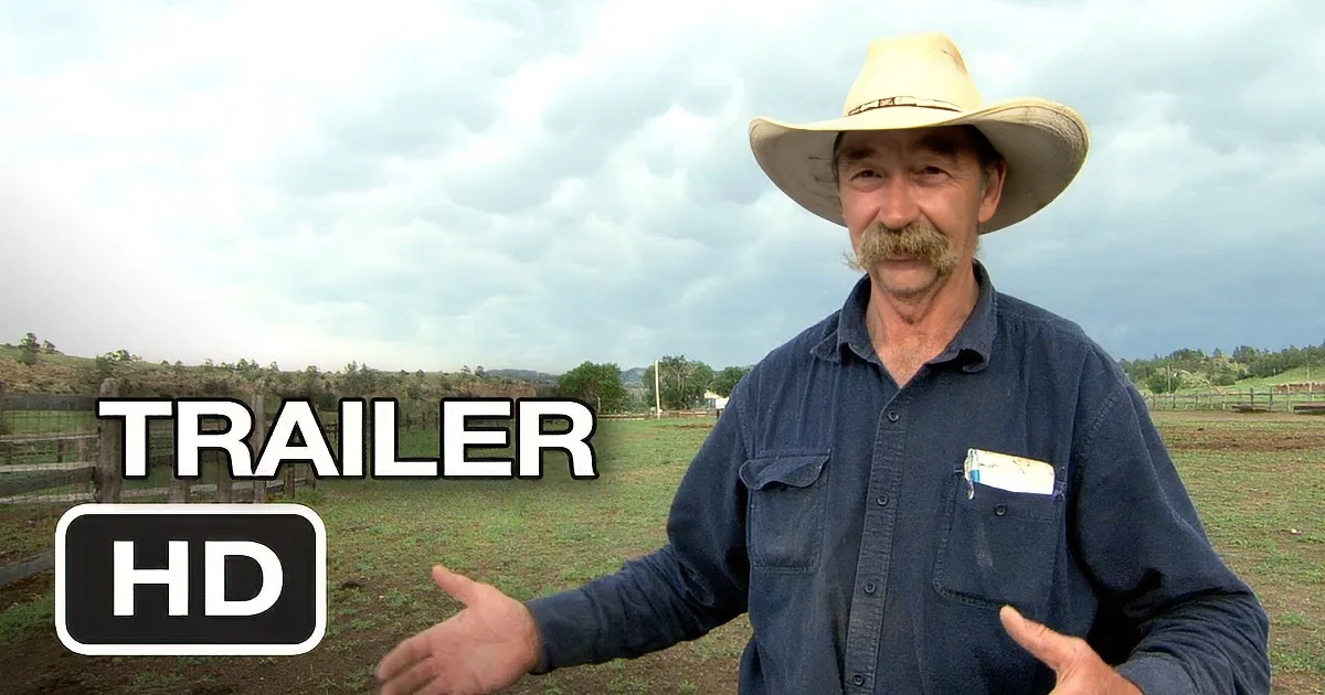 A man in a cowboy hat and blue shirt stands in a field, gesturing with his hands.