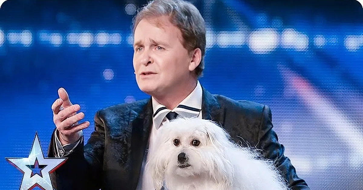 A man in a suit gestures while holding a fluffy white dog on stage.