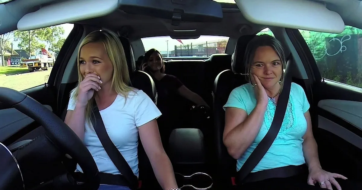 Two women sit in a car, one in the driver's seat, both wearing seatbelts, with a third person in the back seat.