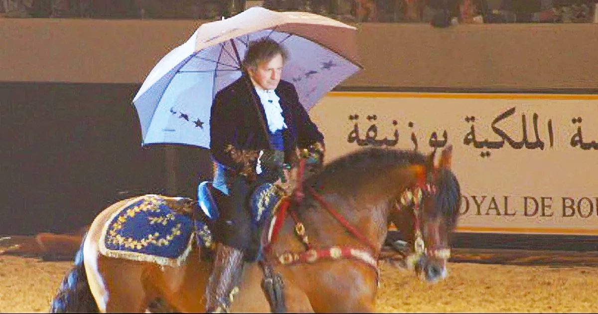 A man in formal attire rides a horse under an umbrella in an arena.