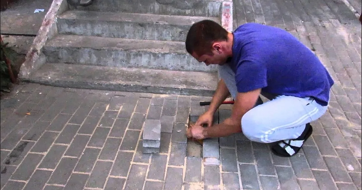 A man in a blue shirt and white pants is kneeling and working on a brick pavement, using a trowel to lay bricks.