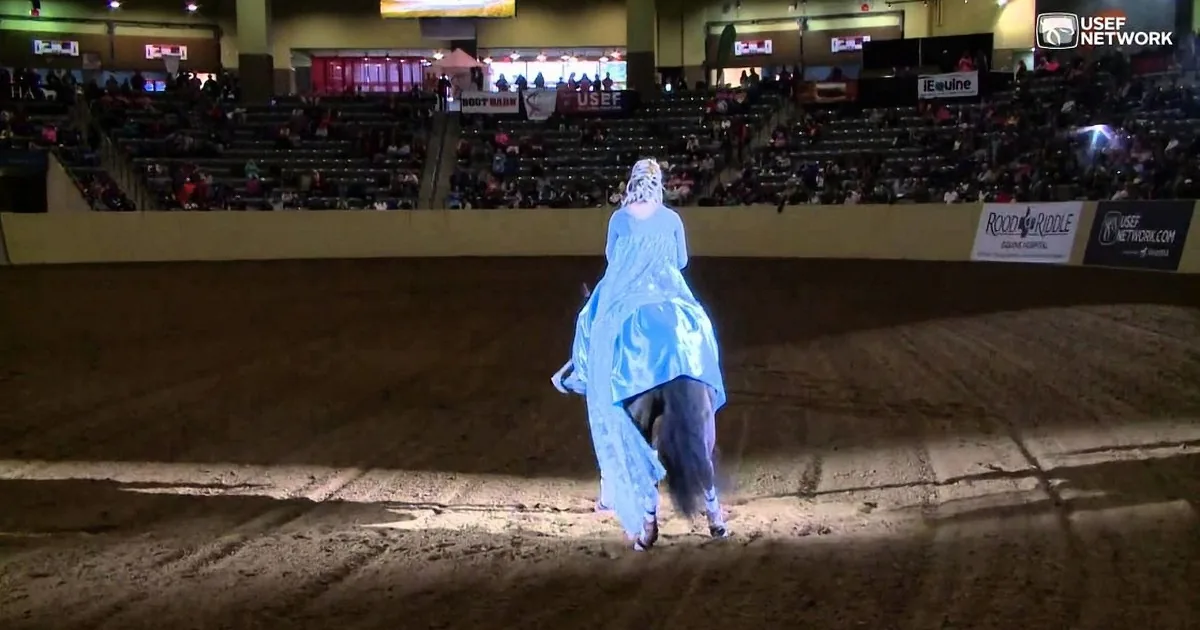 A person in a blue dress rides a horse in an arena with spectators and advertisements in the background.