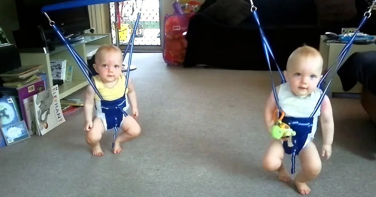 A baby wearing a harness and holding a toy is squatting on a carpeted floor.