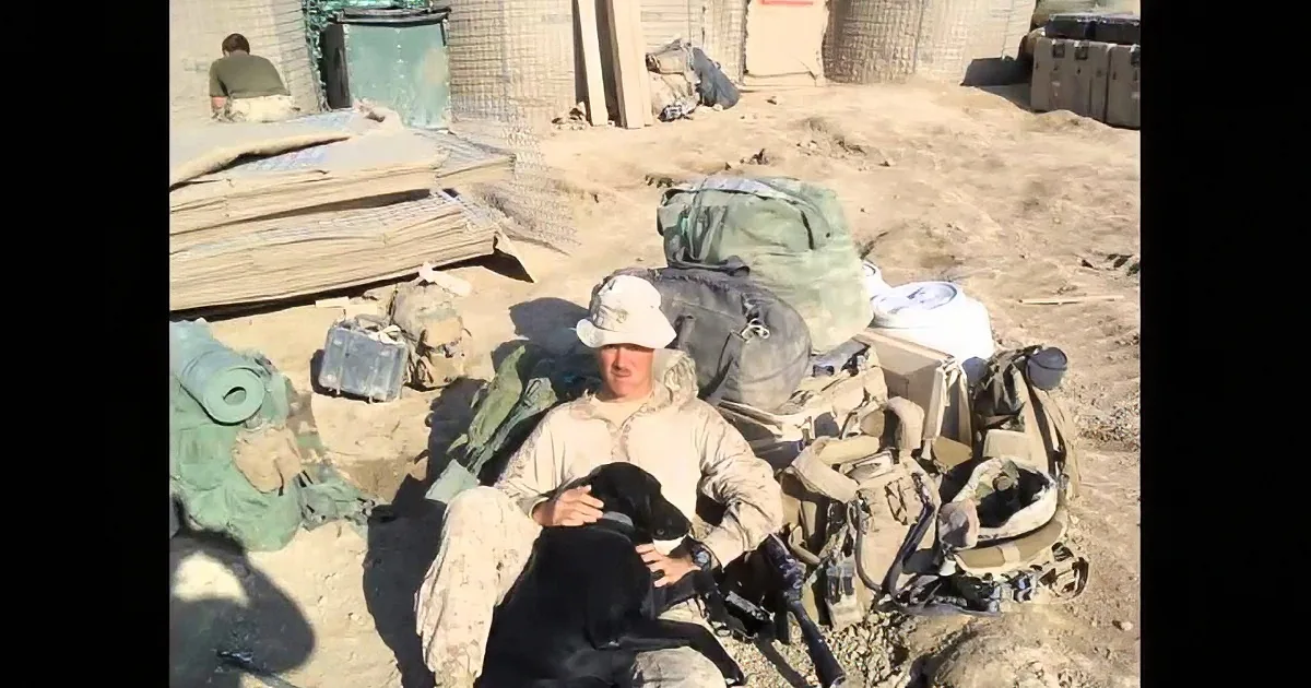 A soldier in a desert uniform sits surrounded by gear and sandbags, holding a dog.
