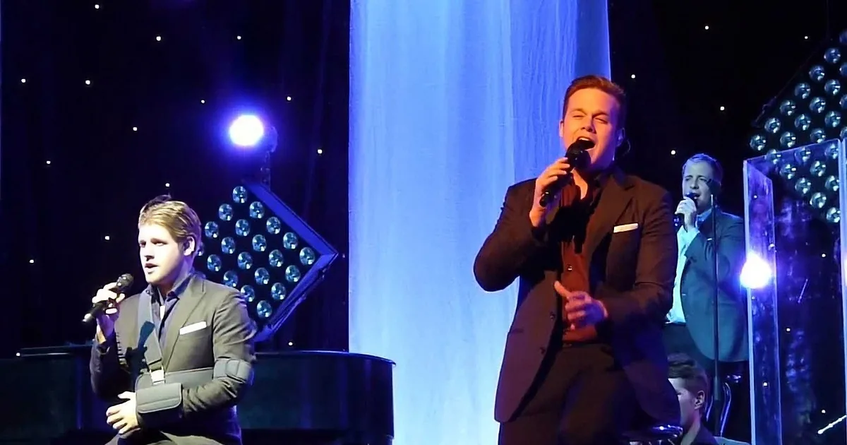 A man in a suit sings into a microphone on stage with other musicians.