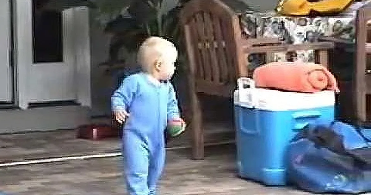A baby in blue pajamas walks outside near a blue cooler and a chair.