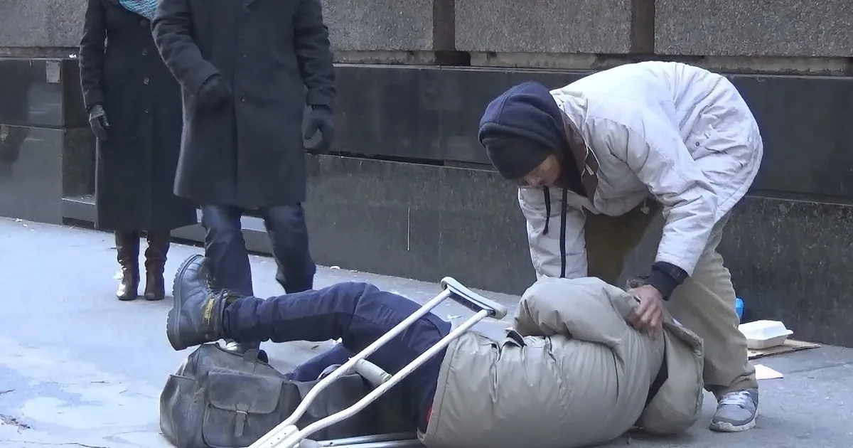 A person in a white jacket helps another lying on the ground with crutches, while others watch.