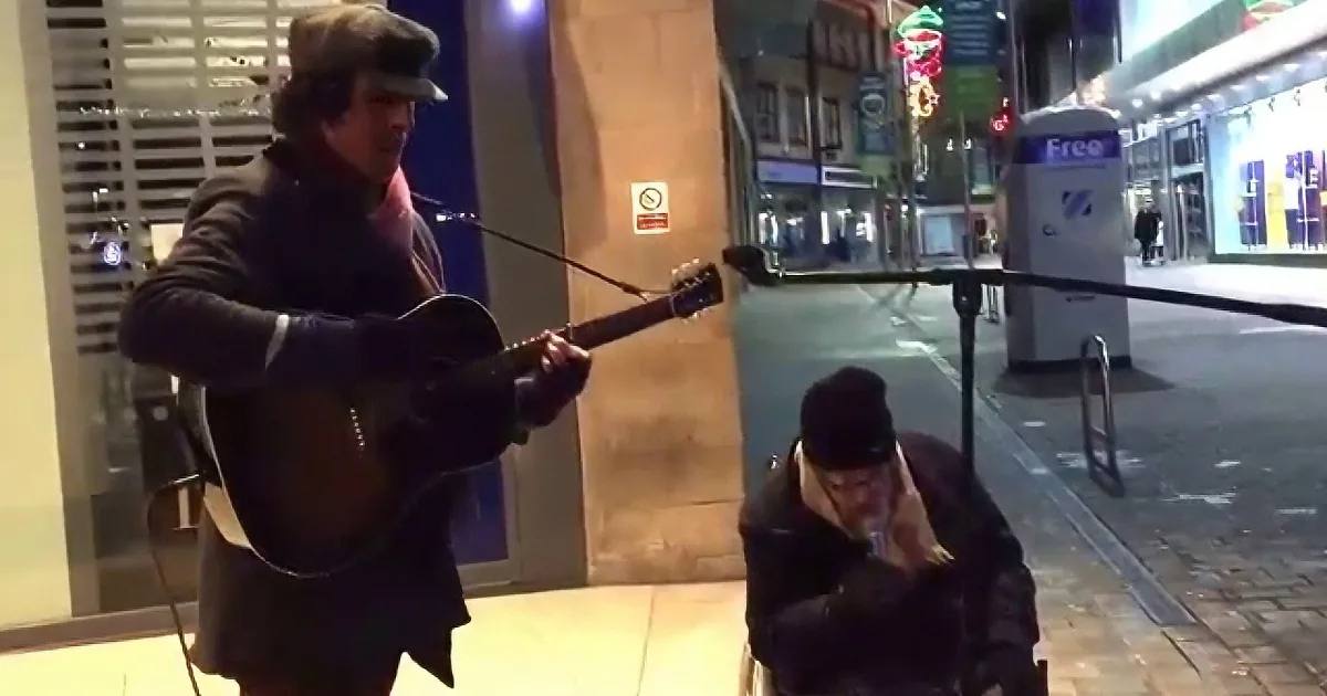 A man in a hat plays guitar on a city street, while another man sits nearby, listening.