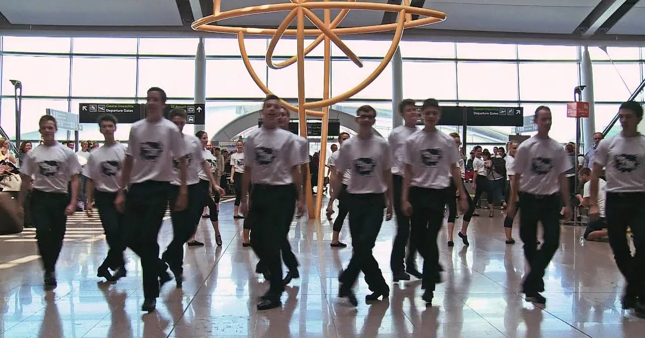 A group of dancers in white shirts and black pants perform in a brightly lit airport terminal.