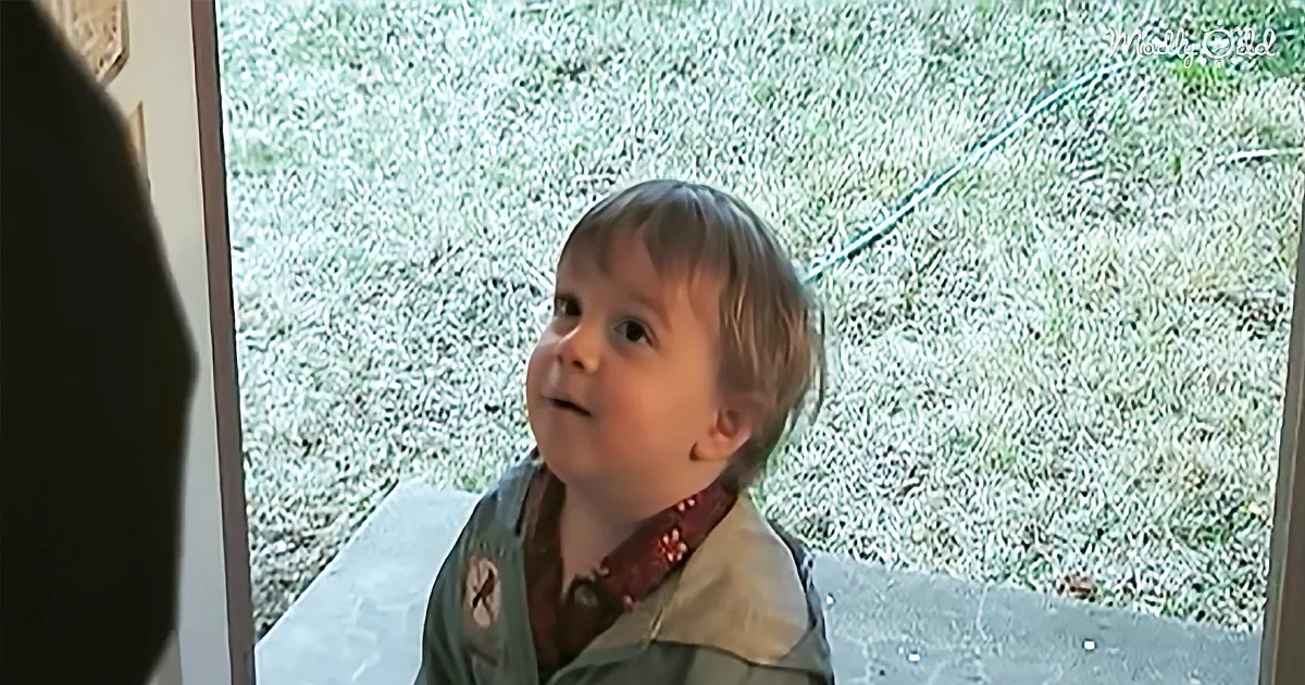 A young child with short hair, wearing a green shirt, looks up with a curious expression, standing on a concrete surface.