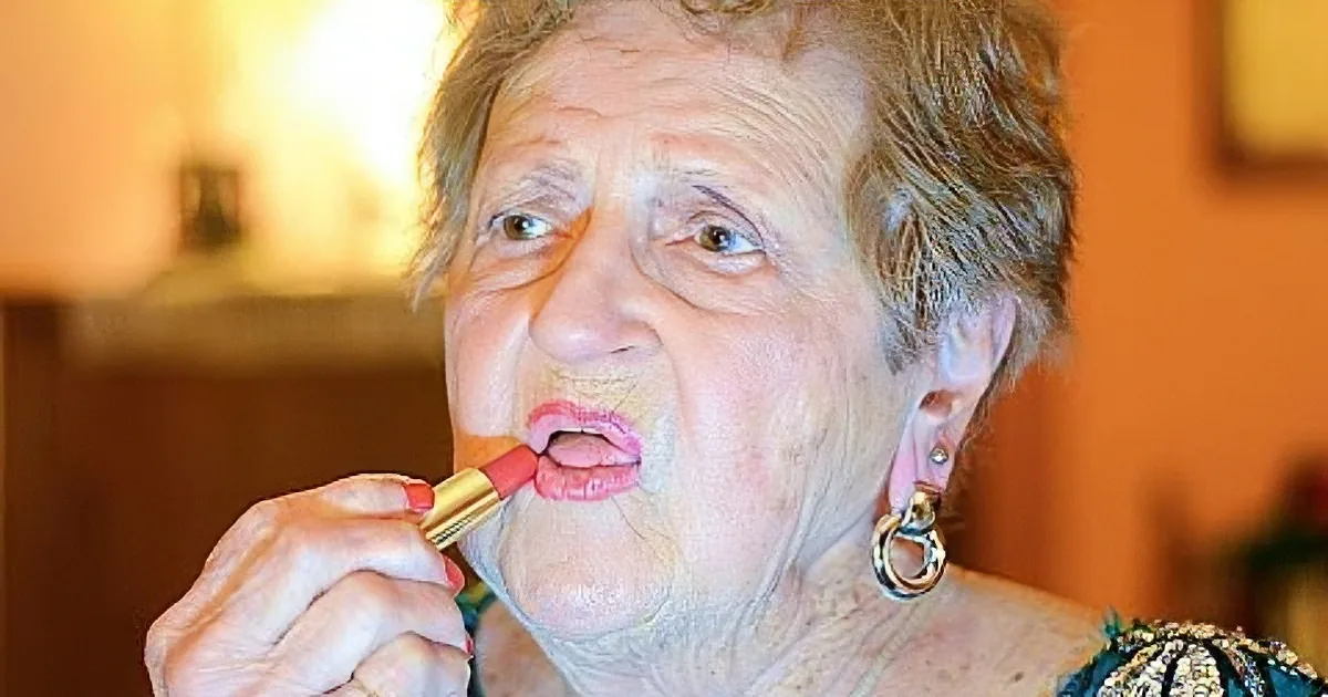 An elderly woman applies pink lipstick, showcasing her earrings and patterned top.