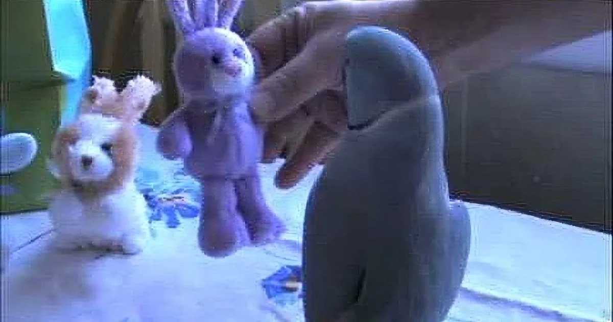 A hand holds a purple stuffed rabbit, surrounded by other stuffed animals on a table.