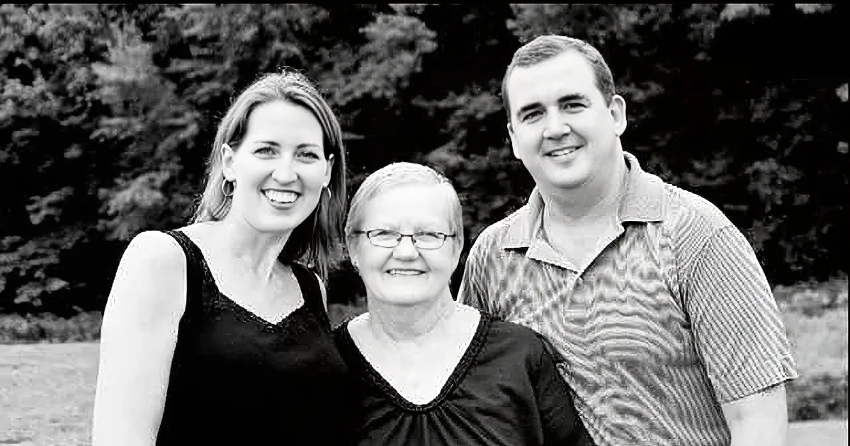 A black-and-white photo shows a woman smiling with two others, one wearing glasses, in a natural setting.