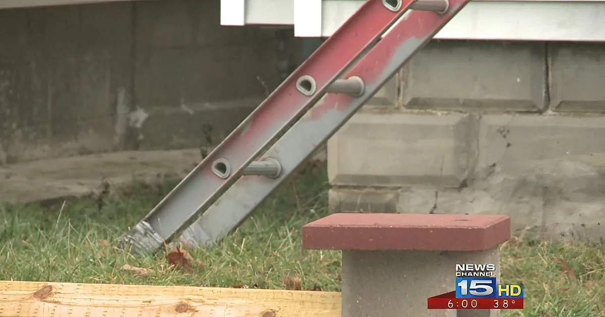 A red ladder leans against a concrete wall, with grass and a wooden plank in the foreground.