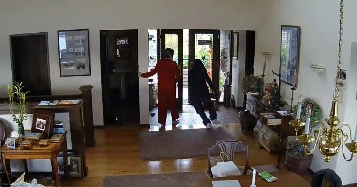 A person in red walks through a doorway into a living room with wooden floors and various furniture.