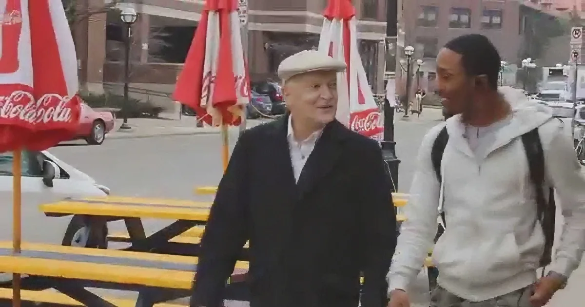 An older man in a white hat and dark coat walks alongside a younger man in a white hoodie and backpack near outdoor...