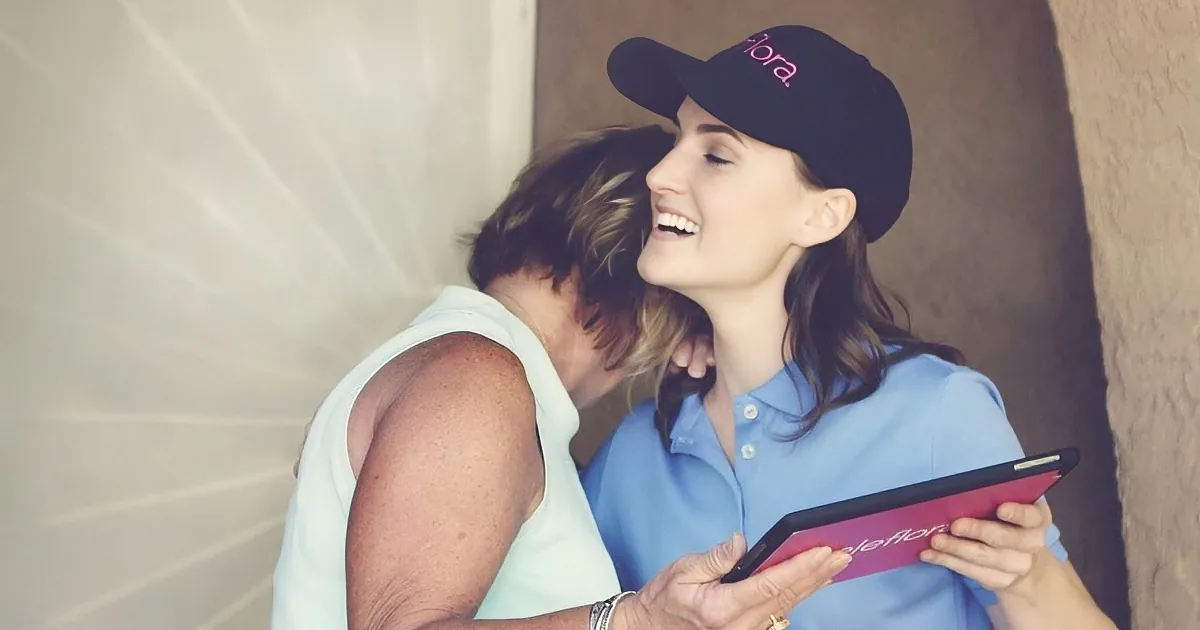 A woman in a blue shirt and black cap holds a tablet, smiling while another woman hugs her from behind.