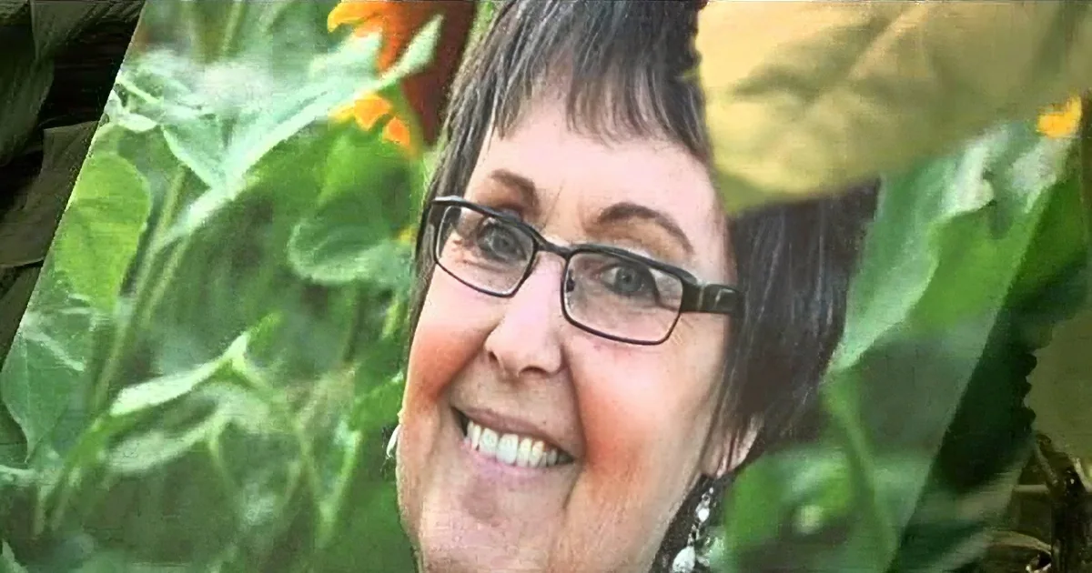 A smiling woman with glasses, surrounded by green foliage and flowers.