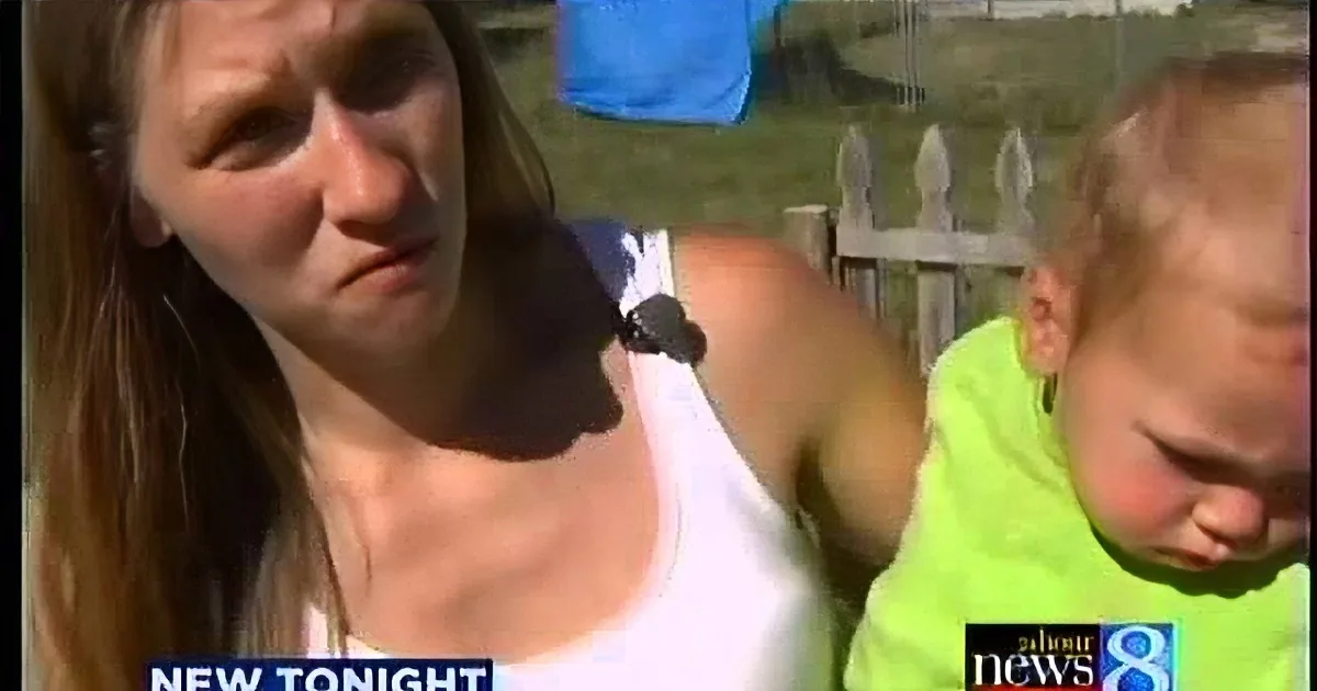 A woman in a white tank top is speaking on a news broadcast, with a baby in a green shirt and a blue tarp in the background.