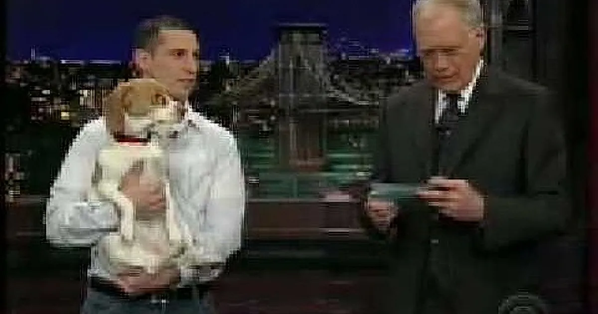 A man holds a small dog while another man in a suit looks on, both seated on a talk show set with a cityscape backdrop.