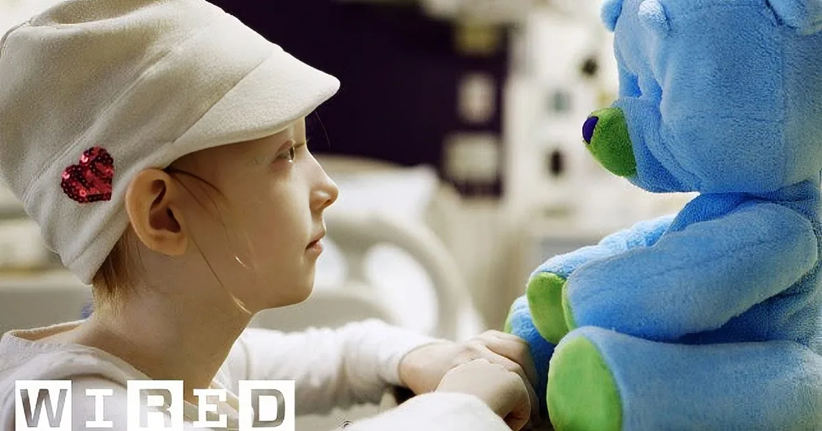 A child wearing a white hat with a red heart, looking at a blue teddy bear in a hospital setting.