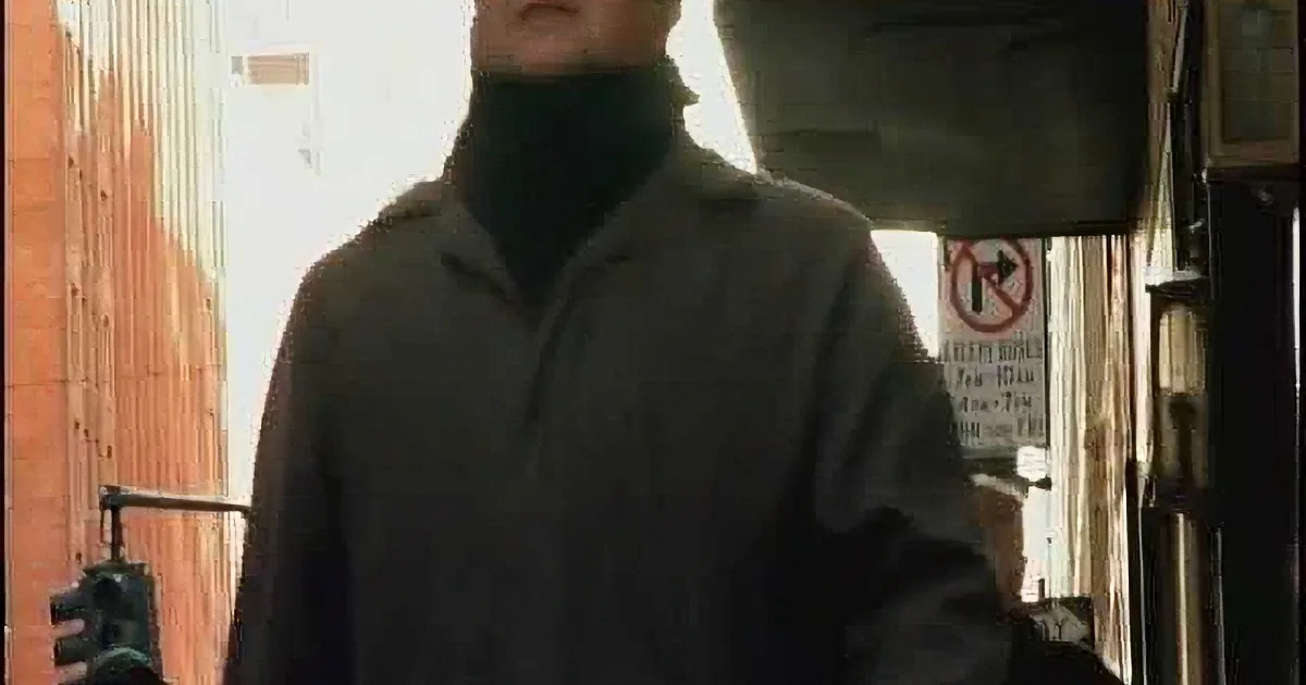 A man in a dark jacket and turtleneck stands in a narrow alleyway, holding a black object.