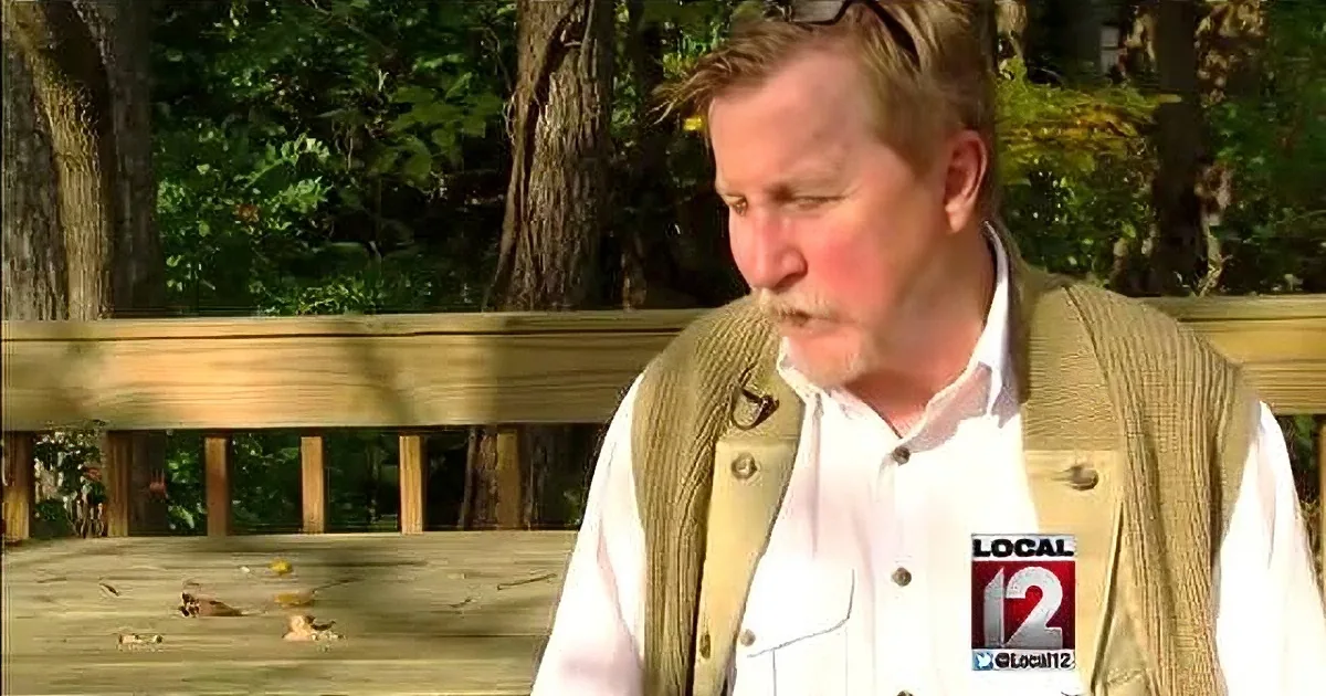 A man in a white shirt and beige vest sits on a wooden bench outdoors, surrounded by greenery.