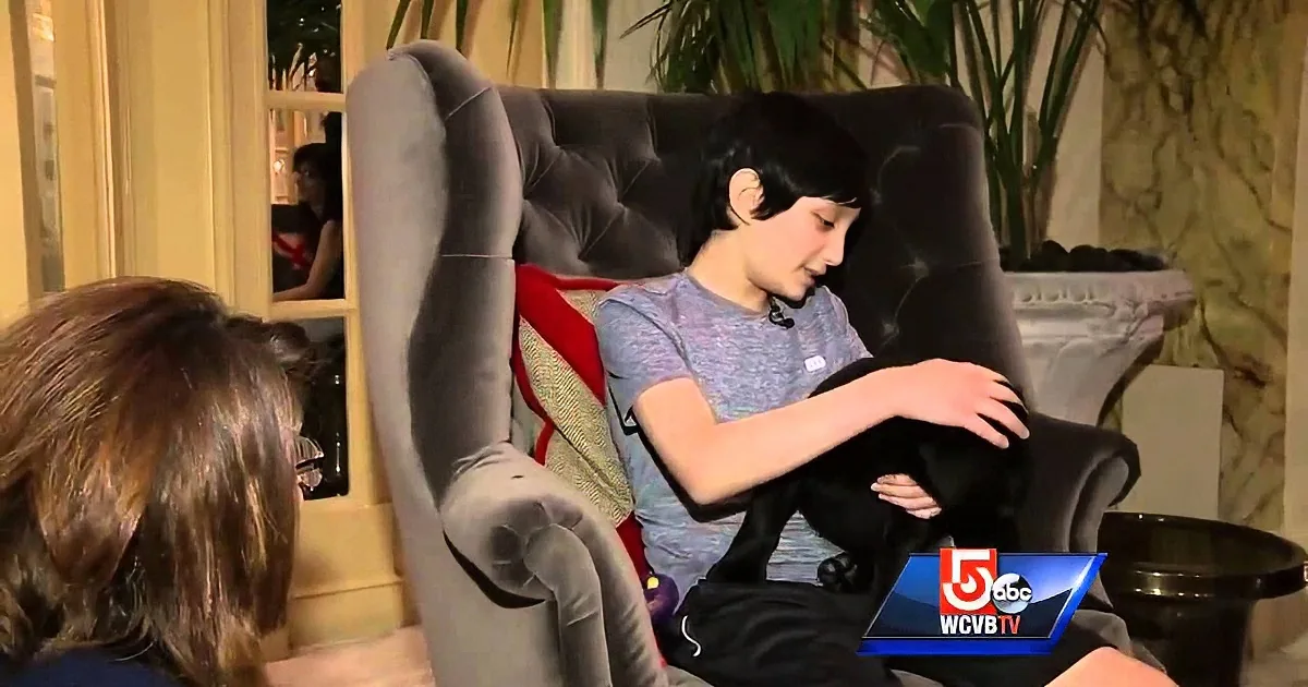 A young boy sits in a chair, adjusting his pants, with a woman in the foreground and a TV screen in the bottom right corner.