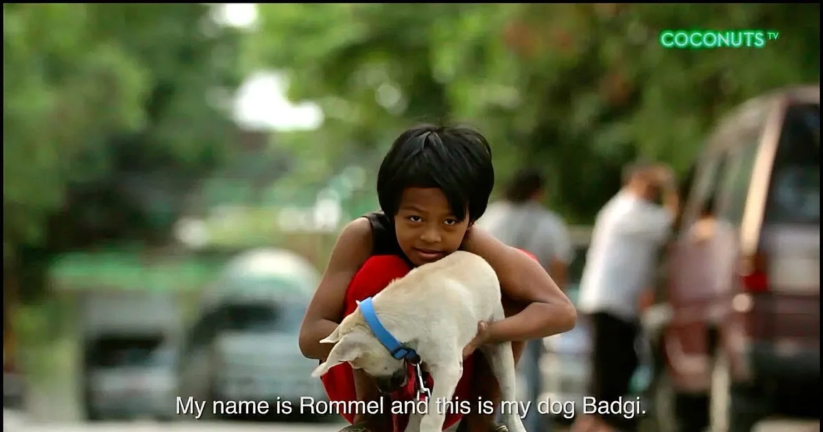 A child named Rommel rides a motorcycle with his dog Badgi, both wearing red and blue harnesses.