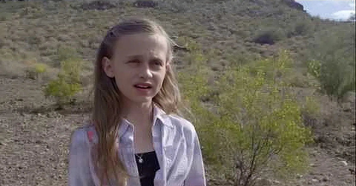 A young girl with long blonde hair stands in a desert-like environment, wearing a white shirt over a black top.