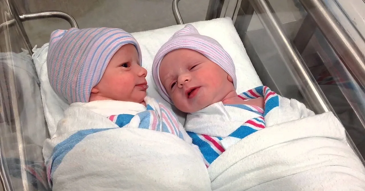 Two newborn babies wrapped in blankets and wearing striped hats lie side by side in a hospital bassinet.