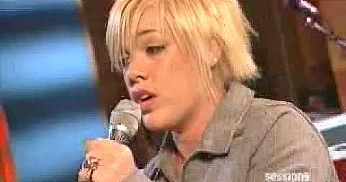 A person with short blonde hair holds a microphone, wearing a brown jacket, in a close-up shot.