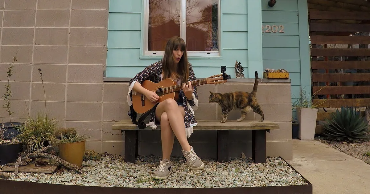 A woman plays guitar on a bench, a cat jumps nearby, in front of a house with a gravel garden.