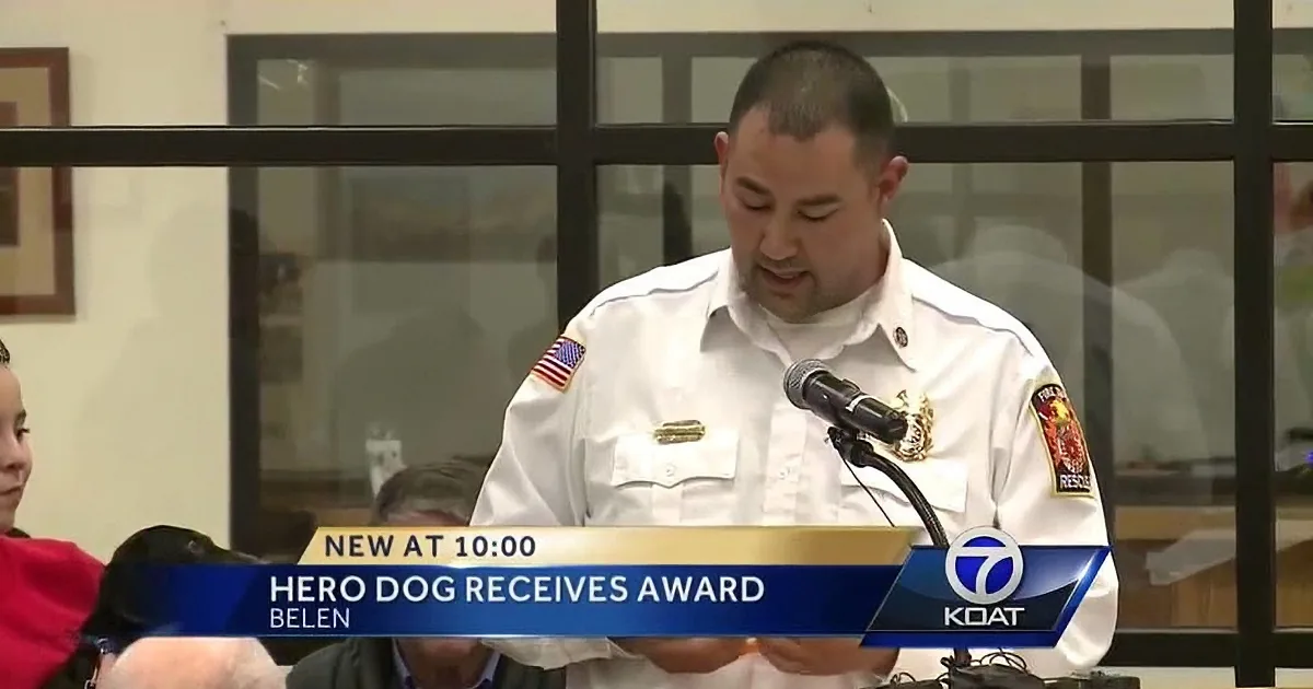 A man in a white uniform speaks into a microphone at a podium, with text indicating a "Hero Dog Receives Award."