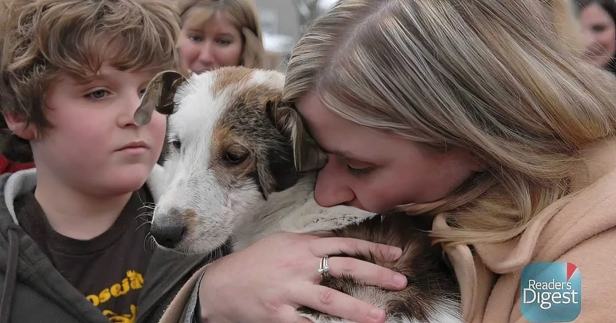 A woman hugs a dog while a boy watches, both smiling, with a crowd in the background.