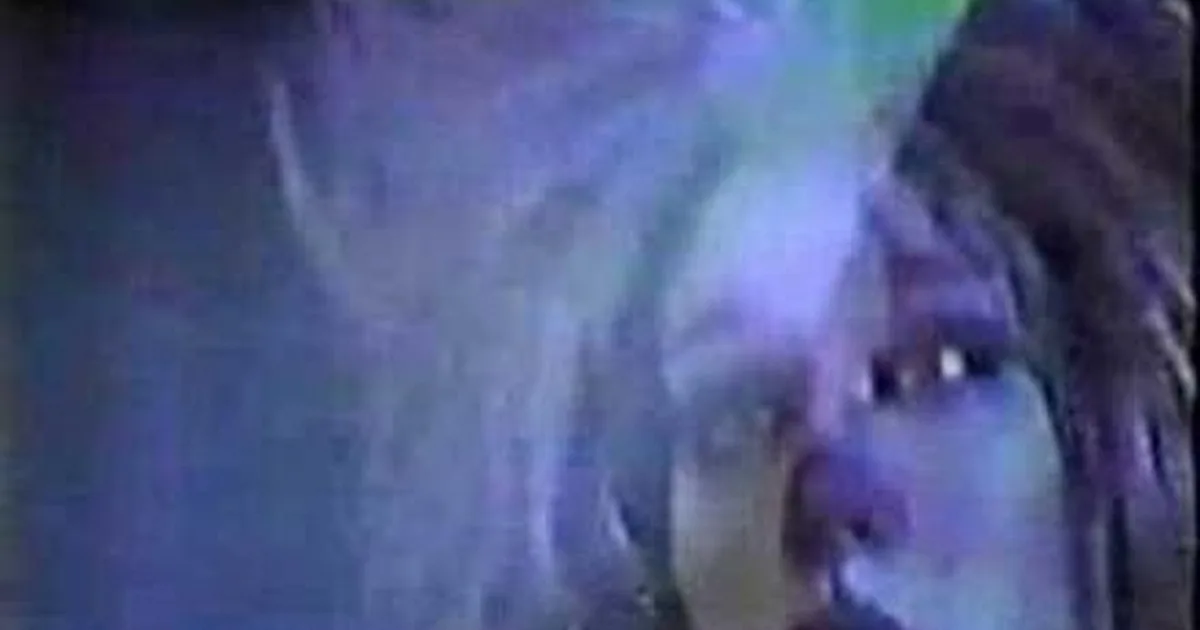 A person with dreadlocks and a blue-toned face is shown in a close-up, possibly in a dimly lit environment.